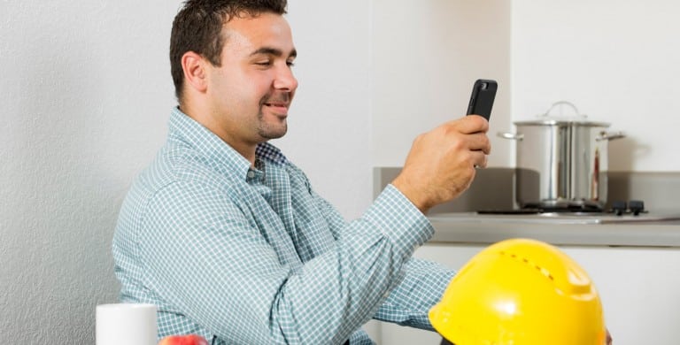 A man in a blue checkered shirt sits next to his yellow hard hat while checking messages on his cell phone.