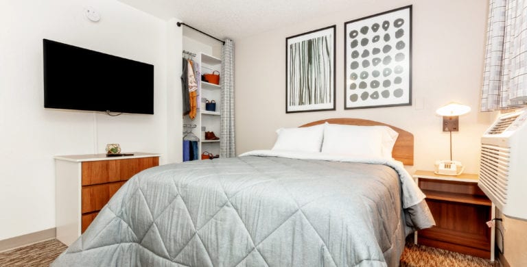 A room with a bed in the middle of the room. The bedspread is grey, and it has 2 white pillows. There are two frames with black lines and circles over the bed. There is a TV on the side wall next to an open concept closet.