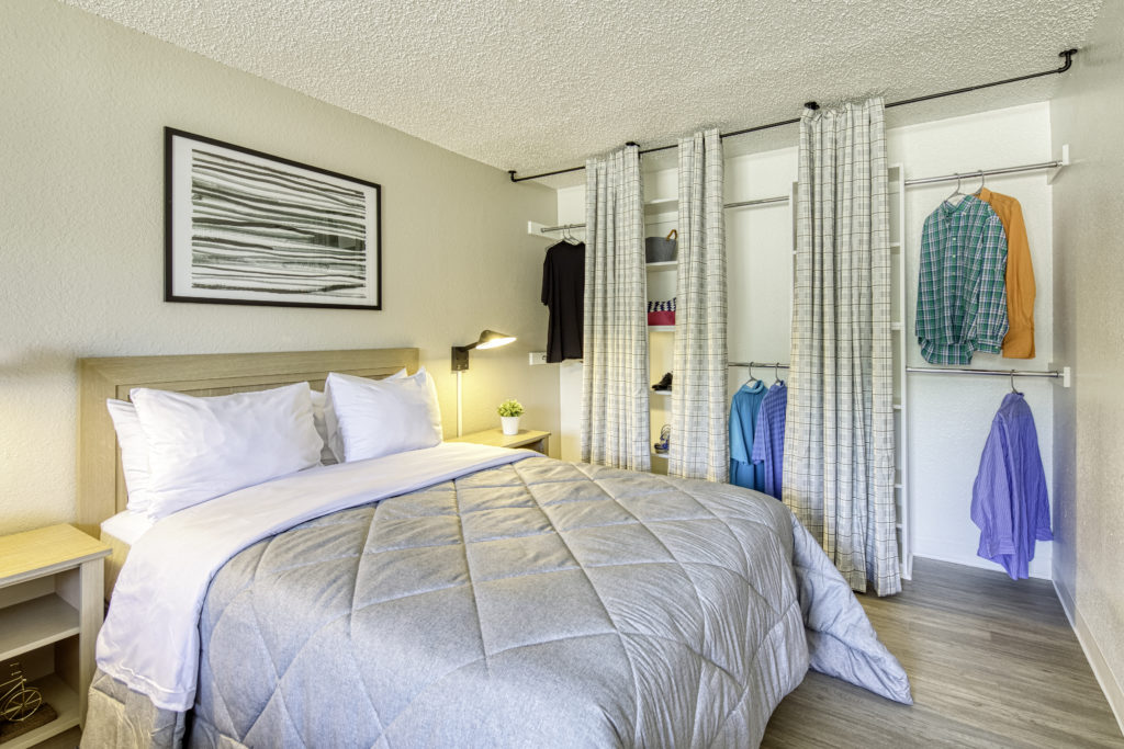 A suite with a bed in the middle. The bed has a hrey comforter and a black and white painting over it. There is an open closet next to the bed.
