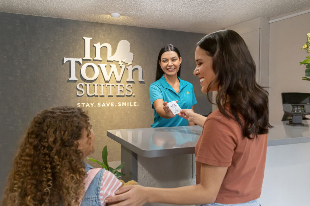 Receptionist in teal uniform hands a card to a guest at a front desk; wall displays 'In Town Suites' behind them.